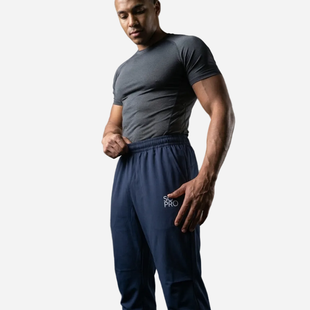 A man in a gym wearing dark gray athletic t-shirt and navy blue sweatpants with a white 'SOPRO' logo on the thigh, posing in front of a concrete wall and weight rack.